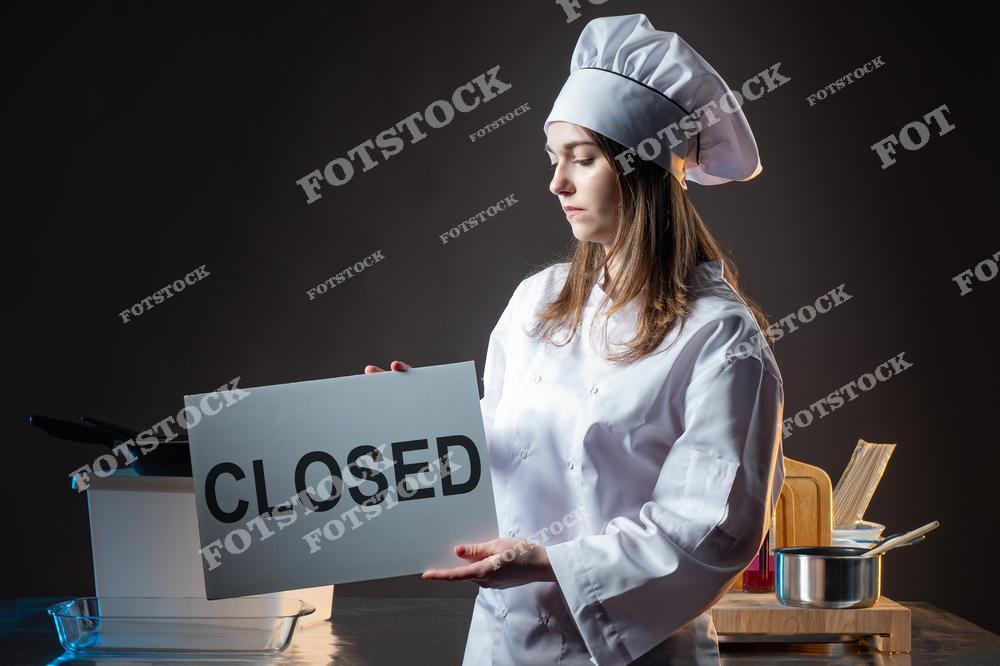 Женщина в форме повара держит табличку с надписью CLOSED. Она стоит перед прилавком кафе, на котором разложены кухонные принадлежности и книги. Обстановка темная, освещена направленным светом, подчеркивающим серьезное выражение лица женщины.